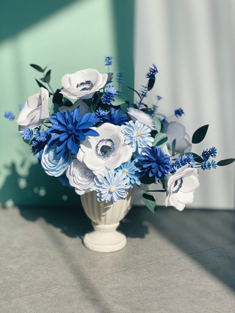 A professional paper flower bouquet featuring a gradient of blue cardstock flowers, including royal blue, sky blue, and navy, mixed with elegant white anemones in a classic cream vase.