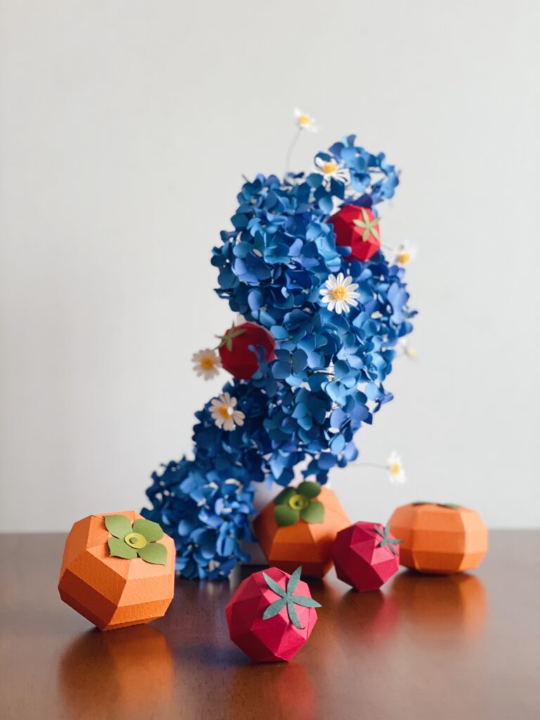 A sophisticated paper art arrangement featuring vibrant blue hydrangeas made of cardstock, accented with small white daisies and red geometric fruit models, showing a bold complementary color contrast.