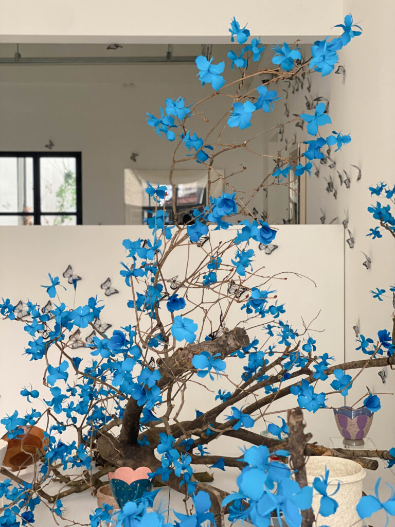 A large-scale paper art installation by Seulpaper, featuring vibrant blue paper flowers blooming on dark brown wooden branches with black paper butterflies on the background, inspired by the movie Avatar.